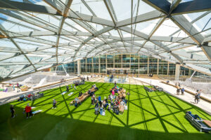 People sitting and relaxing on benches and artificial grass inside a modern glass dome structure.