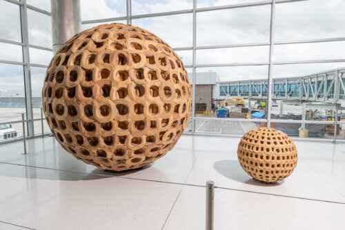Large and small wooden spherical sculptures with cutouts, displayed in a modern airport terminal.
